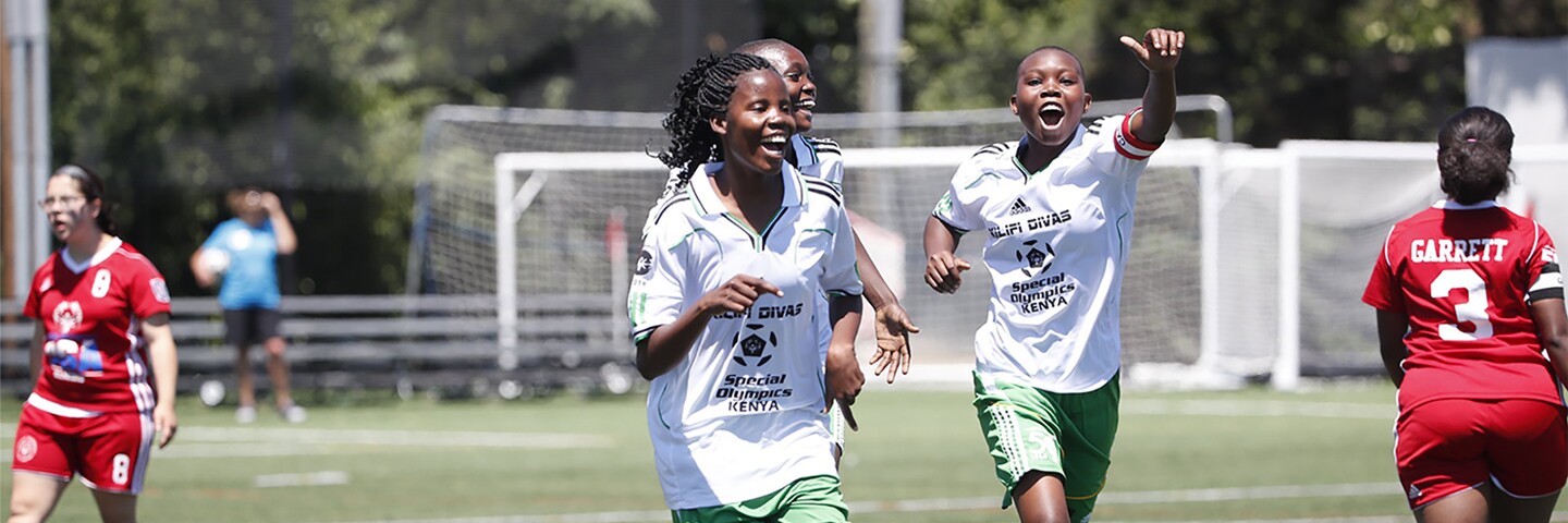 Three girls in white and green on the pitch celebrating and two girls in red. 