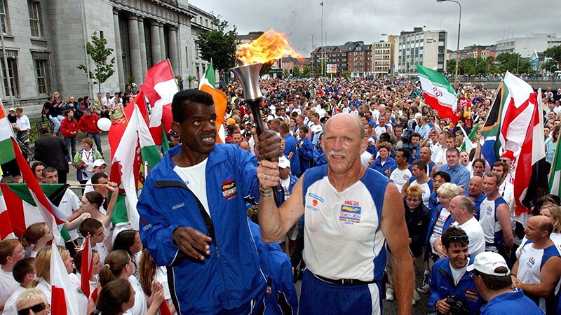 Two runners holding the torch as hundreds of spectators watch. 