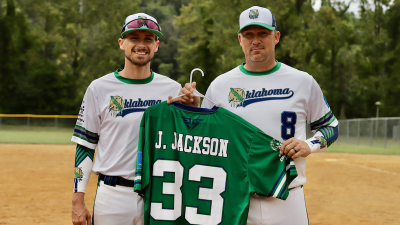 Two men stand on a softball field holding a green softball jersey. 