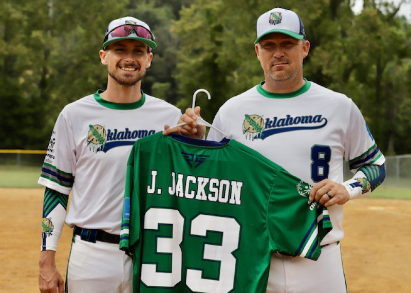 Two men stand on a softball field holding a green softball jersey. 