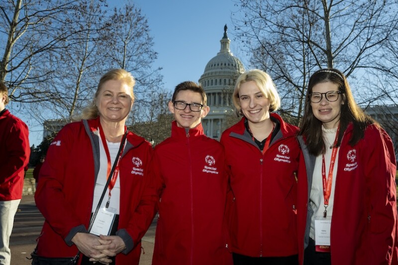 Four people (3 women, 1 man) stand in front of the US Capitol for a photo. All are wearing red jackets with the Special Olympics logo on them. 