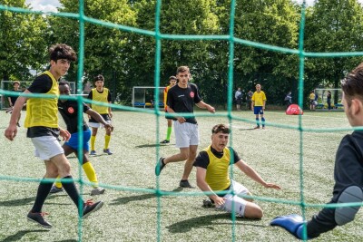 A group of young men in t-shirts, shorts and bibs in action facing the goal on a football pitch.