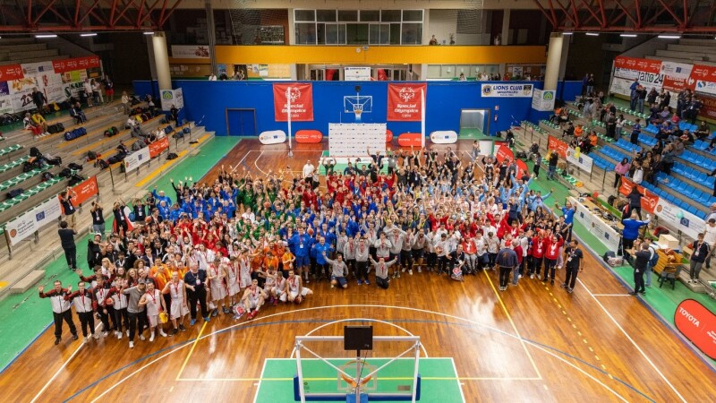 group shot of about 250 people on an indoor basketball court. All standing with their arms in the air. They are wearing colourful tracksuits.