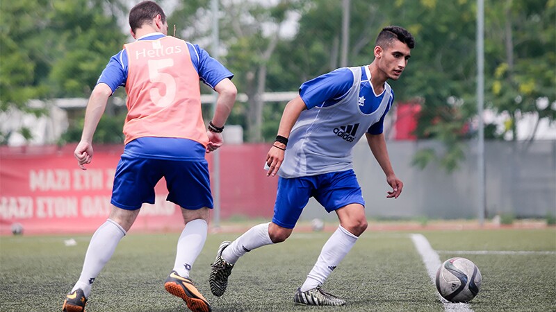 Two footballer players in blue kits chasing a football on an astroturf football pitch. 