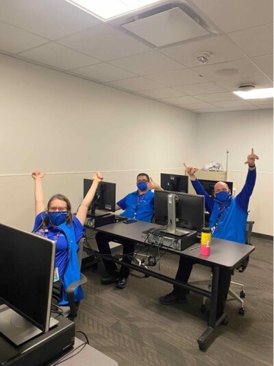 A group of people sit at computers and raise their arms for a picture. 