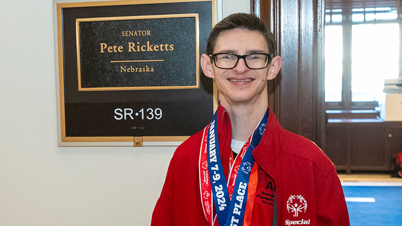 Wyatt Spalding stands in front of a sign for Senator Ricketts office. 