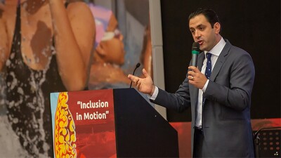 Alt: A man in a suit holding a microphone stands behind a podium branded with the words ‘Inclusion in Motion’ and a large poster of a swimmer with hands grasped in celebration is beside him.