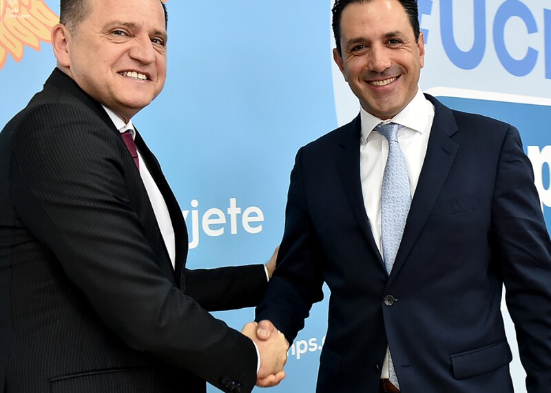 Two men wearing suits, shake hands and smile at the camera