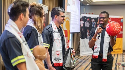 yasha Derera, Special Olympics athlete leader from Zimbabwe holding a microphone, presents a Play Unified red ball and ceremonial scarves to four Medi Response first aid volunteers in paramedic uniforms on the stage of the conference.