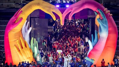 A tunnel is full of delegations entering the stadium as they walk under a huge sculpture of colorful hands creating a heart shape.