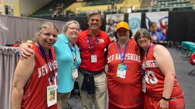 Four women and a man pose for a photo. The women have red basketball uniforms on and everyone has their arms around each other. 