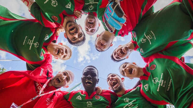 Footballers form the Special Olympics Italia team standing in a circle looking down at the camera and the camera shooting the photo looking up; the sky is in the background. 