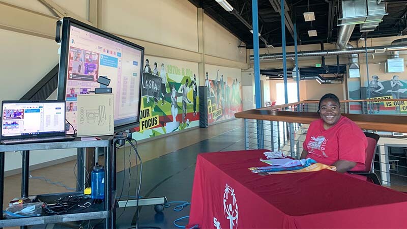 A Special Olympics athlete sits behind a table covered in a red cloth. In front of her are video cameras and computer monitors. She smiles at the camera. 