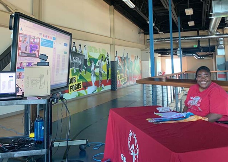 A Special Olympics athlete sits behind a table covered in a red cloth. In front of her are video cameras and computer monitors. She smiles at the camera. 