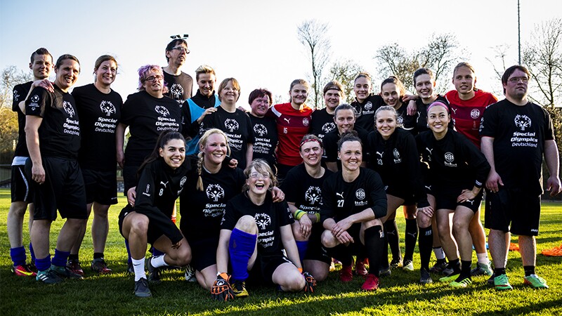 A large group of footballers wearing black Special Olympics t-shirts pose for the camera on a grass pitch. 