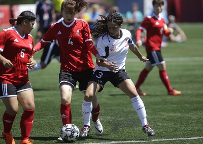 Young adult women on the pitch; one player from the opposing team attempts to take the ball. 