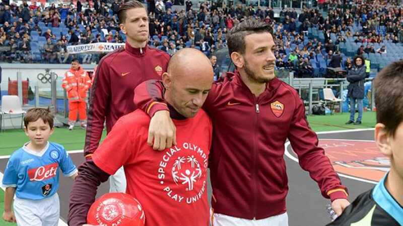 Pro footballer with his arm around a unified palyer that is carrying a football onto the field. The unified player is wearing a red t-shirt that says Special Olympics, Play Unified. 