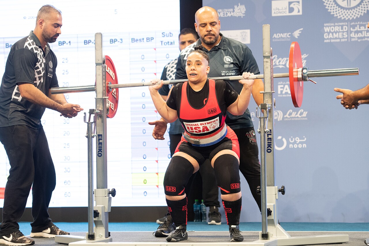 Angel, a power lifter lifting weights over her shoulders. a spotter is on either side of her and two are behind her. 