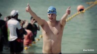 A swimmer with a blue swim cap coming out of the water and giving a thumbs up. 