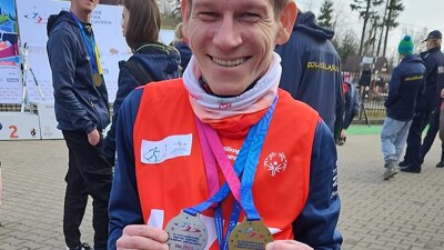 Konrad is wearing a red Special Olympics jersey and smiles at the camera while holding his medals.