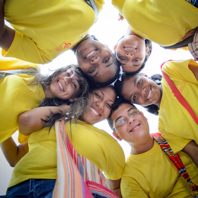 Six young adults in a circle with their heads together looking down and smiling. 