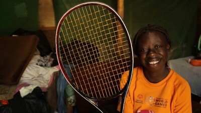 Girl sitting in an orange t-shirt holding a tennis racquet smiling for a photo. 