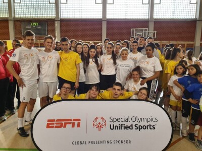 A group of young people in yellow and white t-shirts stand behind an 'ESPN and Special Olympics Unified Sports' banner facing the camera on an indoor court.