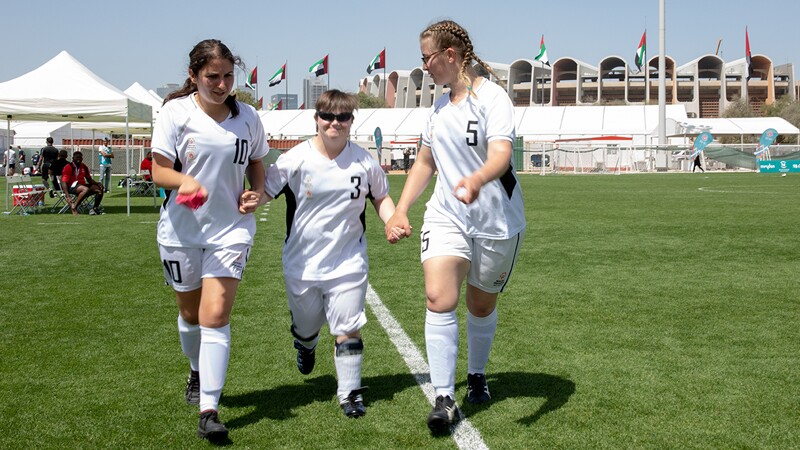 Three female footballers walking on the field hand in hand wearing all white. 