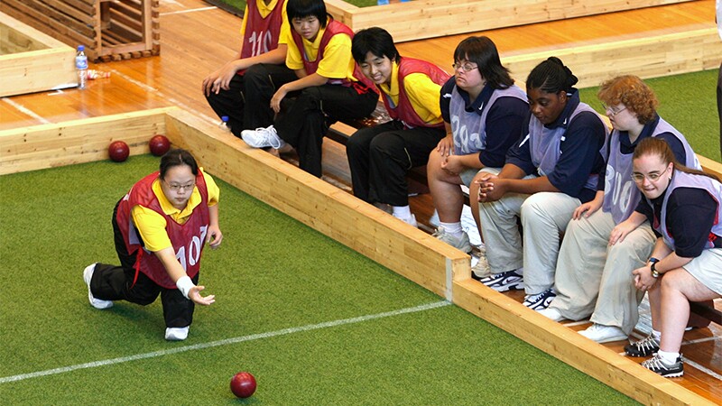 Girl rolling the bocce ball while her team and opposing team on the sidelines watch. 