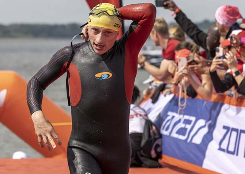 Male competitor runs in wet suit on red carpet with open water in the background. 