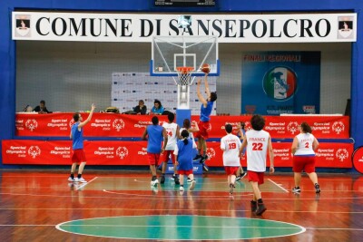 a group of young people - both male and female - wearing basketball uniforms and playing basketball on a court. In the center, a young male athlete is jumping to the hoop with a basketball in his hands, attempting to score.