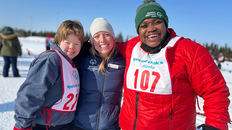 A group of three people pose for a photo. They are outside wearing heavy winter coats and the ground is covered in snow. 