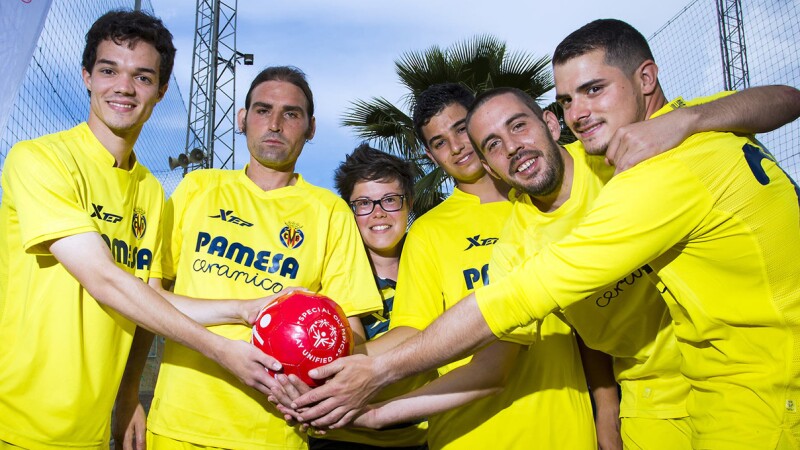 Unified footballer group of 6 people, one female and five male, all wearing yellow shirts and shorts with their hands together holding a red Special Olympics Play Unified football. 