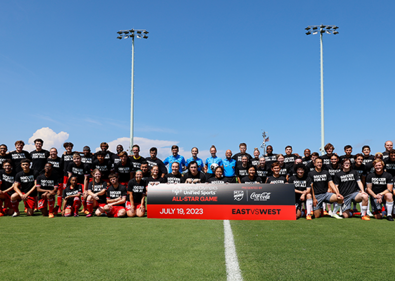 A group of Special Olympics soccer players, coaches and officials pose for a pre-game photo on the soccer field. They're standing behind a sign that says "Special Olympics Olympics Unified Sports All-Star Game". 